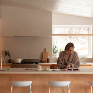 Une personne préparant des aliments dans une cuisine moderne et chaleureuse. L’espace met en valeur des matériaux naturels comme le bois clair et un design épuré. On aperçoit une cuisinière, un four de Our Place, et des accessoires de cuisine soigneusement sélectionnés, créant une atmosphère conviviale et fonctionnelle.