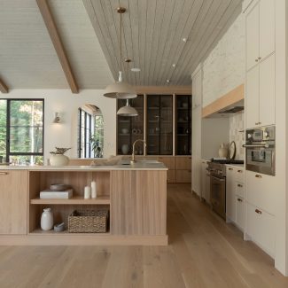 Une cuisine au design chaleureux et naturel, mettant en avant un îlot en bois clair avec des détails en laiton et un plan de travail en pierre claire. Des étagères ouvertes accueillent des paniers tressés et de la vaisselle minimaliste. Le dosseret en carreaux de céramique brillants et le mur en pierre texturée ajoutent du caractère à l’espace, tandis que deux suspensions beiges apportent une touche d’élégance discrète.