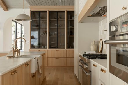 Une cuisine au design chaleureux et naturel, mettant en avant un îlot en bois clair avec des détails en laiton et un plan de travail en pierre claire. Des étagères ouvertes accueillent des paniers tressés et de la vaisselle minimaliste. Le dosseret en carreaux de céramique brillants et le mur en pierre texturée ajoutent du caractère à l’espace, tandis que deux suspensions beiges apportent une touche d’élégance discrète.