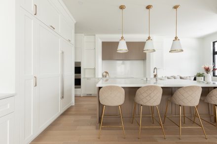 Une cuisine contemporaine avec un grand îlot en quartz blanc, accompagné de tabourets en velours beige aux pieds dorés. Les luminaires suspendus apportent une élégance intemporelle, tandis que les armoires blanches encadrent l’espace avec raffinement.