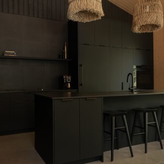 : Vue plus large de la cuisine moderne entièrement noire, avec un îlot fonctionnel et des suspensions en fibres naturelles qui contrastent avec l’esthétique épurée. Un mur en bois clair et une grande fenêtre laissent entrer la lumière naturelle.