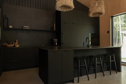  : Vue plus large de la cuisine moderne entièrement noire, avec un îlot fonctionnel et des suspensions en fibres naturelles qui contrastent avec l’esthétique épurée. Un mur en bois clair et une grande fenêtre laissent entrer la lumière naturelle.