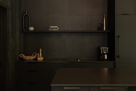 Cuisine moderne entièrement noire, avec un îlot fonctionnel et des suspensions en fibres naturelles qui contrastent avec l’esthétique épurée. Un mur en bois clair et une grande fenêtre laissent entrer la lumière naturelle.