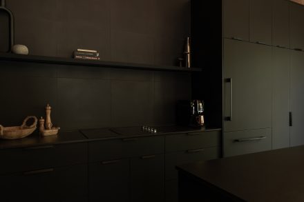 Cuisine moderne entièrement noire, avec un îlot fonctionnel et des suspensions en fibres naturelles qui contrastent avec l’esthétique épurée. Un mur en bois clair et une grande fenêtre laissent entrer la lumière naturelle.