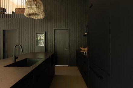 Cuisine moderne entièrement noire, avec un îlot fonctionnel et des suspensions en fibres naturelles qui contrastent avec l’esthétique épurée. Un mur en bois clair et une grande fenêtre laissent entrer la lumière naturelle.